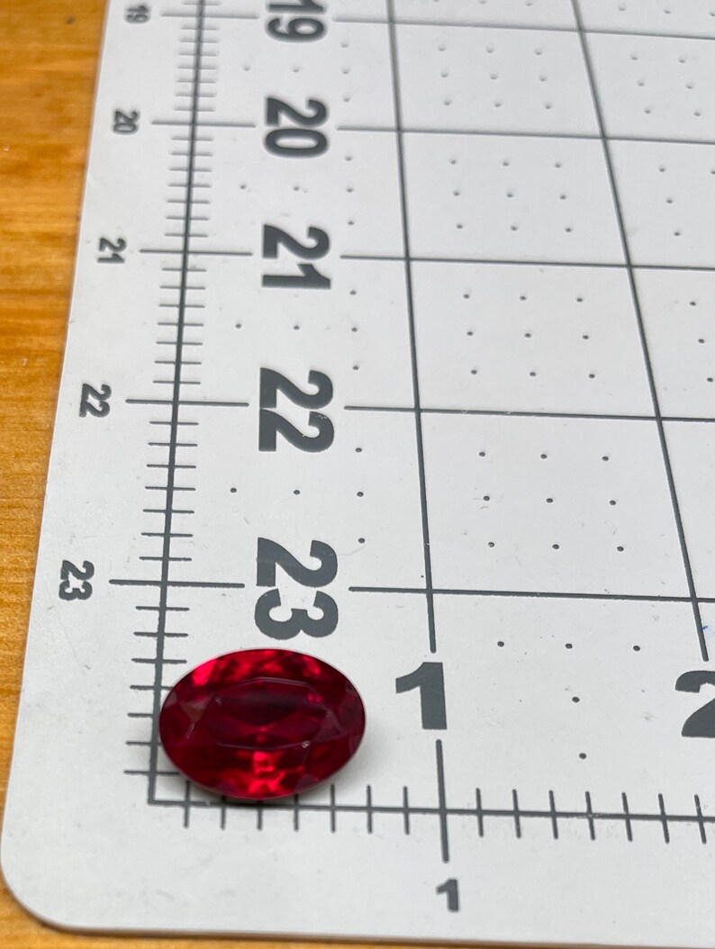 May include: A deep red oval-shaped gemstone measuring approximately 1 inch in length. The gemstone is sitting on a white gridded ruler.