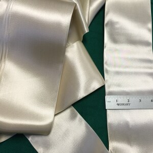 May include: A roll of ivory satin ribbon with a double-stitched edge. The ribbon is 6 inches wide and is measured with a ruler.