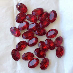 May include: A collection of oval-shaped, red, faceted glass gems. The gems are scattered on a white surface.