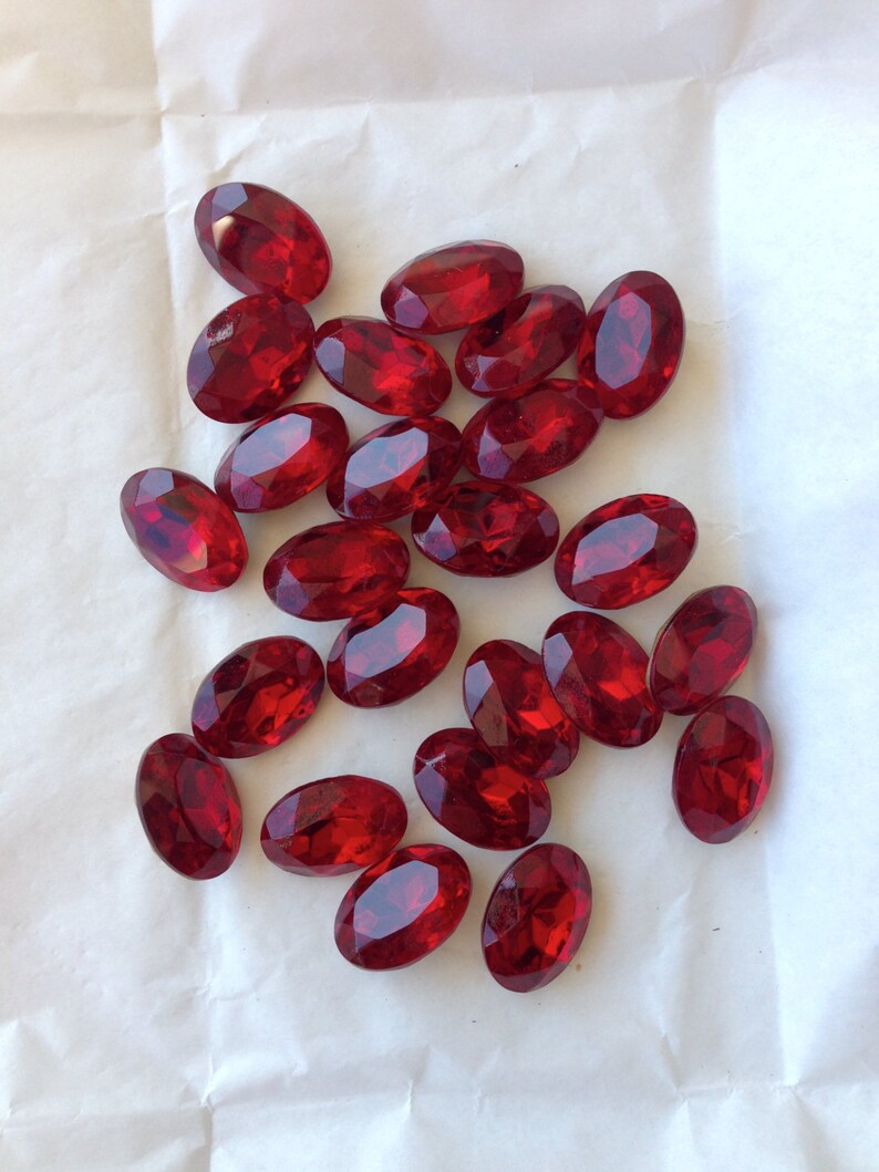 May include: A collection of oval-shaped, red, faceted glass gems. The gems are scattered on a white surface.