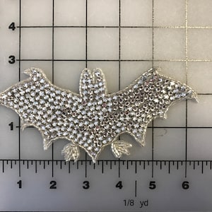 Peut inclure: Un patch thermocollant en forme de chauve-souris argenté scintillant. La chauve-souris mesure environ 10 cm de large et 7,5 cm de haut. Elle est recouverte de petits strass transparents.