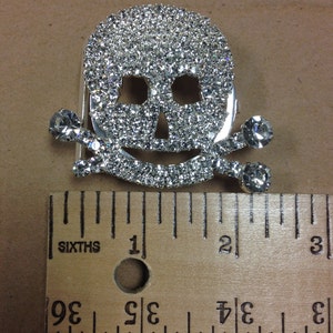 Puede incluir: Un broche de plata con forma de calavera y huesos cruzados con diamantes de imitación. El broche mide aproximadamente 2,5 cm de ancho.
