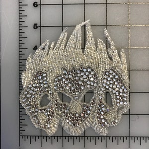 Puede incluir: Una aplicación de calavera de cuentas blancas y pedrería con un borde deshilachado. La aplicación mide aproximadamente 10 cm de ancho y 12,5 cm de alto.
