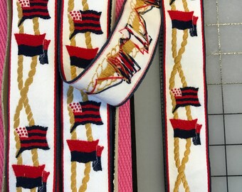 May include: White fabric ribbon with a repeating pattern of gold rope, red and blue American flags, and red and blue squares. The ribbon is approximately 1 inch wide.