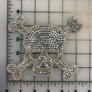 Puede incluir: Un parche de calavera y huesos cruzados plateado cubierto de pedrería transparente. El parche mide aproximadamente 7,6 cm de ancho y 7,6 cm de alto.