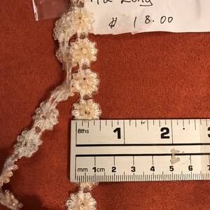 Może przedstawiać: Biała koronka z małymi białymi kwiatami i perłami. Koronka ma 42 cale długości i kosztuje 18,00 €.