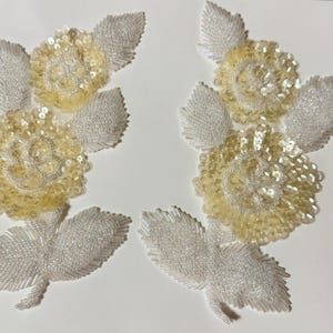 Könnte beinhalten: Zwei weiße und gelbe Blumenapplikationen mit Perlenblättern und Blütenmitten. Die Applikationen bestehen aus Pailletten und Perlen.