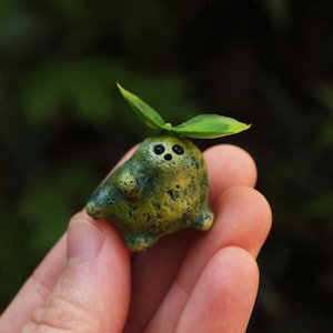 May include: A small, textured, olive-green clay figurine with a mottled surface, featuring two tiny black eyes and a small mouth. A single green leaf sprout is attached to the top of the rounded, bulbous body, which has two short legs.