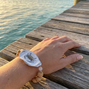 May include: A bracelet featuring a painted crab design on a white shell, connected to wooden beads and a pink gemstone. The bracelet is displayed on a wooden surface with a serene ocean backdrop.