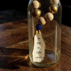 May include: A necklace with wooden beads in natural and gold tones, displayed in front of a clear glass bottle. The necklace has a pendant with the text "We Rise By Lifting Others". The pendant is white with gold edges.