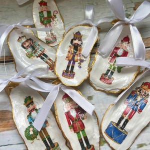 May include: Hand-painted oyster shell Christmas ornaments featuring colorful nutcracker designs. Each shell is edged with gold paint and has a white ribbon for hanging. The ornaments showcase various nutcracker figures in festive attire, perfect for holiday decor.