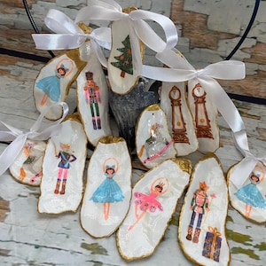 May include: Hand-painted oyster shell ornaments with gold edges and white ribbon. The ornaments feature images of ballerinas, nutcrackers, a Christmas tree, and a rocking horse. These unique ornaments are perfect for holiday decorating.