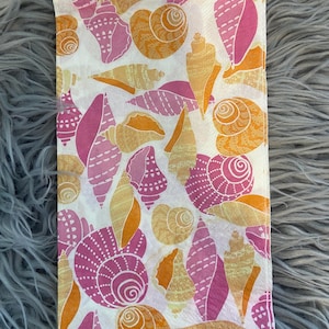 May include: A folded white cloth napkin with a repeating pattern of seashells in shades of pink, orange, and tan. The seashells vary in size and shape, creating a visually appealing design. The napkin is likely made of a soft fabric.