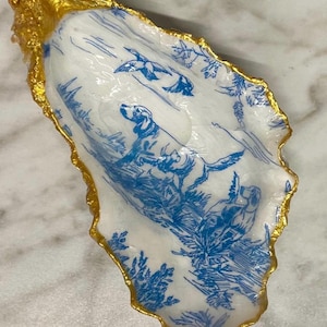 May include: A decorative oyster shell dish with a blue and white hunting scene design. The rim is edged with gold. The dish features a dog, ducks, and foliage.