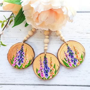 May include: Three circular wood slices hanging from natural twine with wooden beads, each hand-painted with purple, pink, and orange foxglove flowers and green leaves. The word "mint" is written in cursive on the right side of each wood slice.