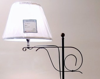 May include: A black metal floor lamp with a white lampshade. The lamp has a curved design and a decorative base. The lampshade has a label attached. The lamp is on a patterned rug.
