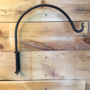 May include: Black metal wall-mounted hook with a curved arm and a decorative scroll at the end. The hook is mounted on a wooden wall.