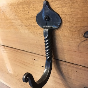 May include: A black metal hook with a twisted stem and a decorative leaf-shaped top. The hook is mounted on a wooden surface.