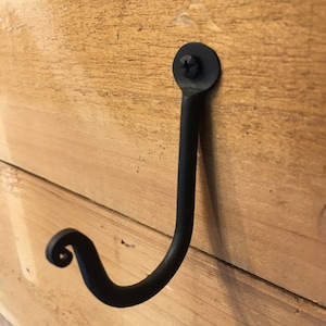 May include: Black metal hook with a decorative scroll at the end. The hook is mounted on a wooden surface.