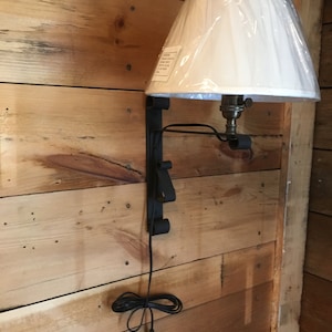 May include: A white lampshade with a black cord and a metal wall mount. The lamp is mounted on a wooden wall.