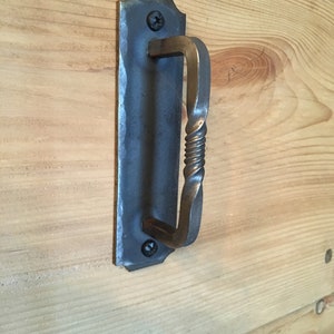 May include: A black metal door handle with a twisted design. The handle is mounted on a wooden surface.