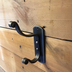 May include: A black metal wall hook with a decorative scroll design at the ends. The hook is mounted on a wooden wall.