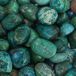Puede incluir: Primer plano de un montón de piedras de crisocola pulidas. Las piedras varían en tamaño y forma, mostrando una mezcla de tonos verde azulado, turquesa y verde oscuro con patrones marrones y azules. Las piedras lisas y redondeadas están dispuestas juntas.