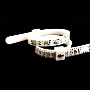 Può includere: Un misuratore di dimensioni dell'anello in plastica bianca con il testo "SIZE & HALF SIZES 1 2 3 4" e i numeri da 7 a 17 stampati su di esso.