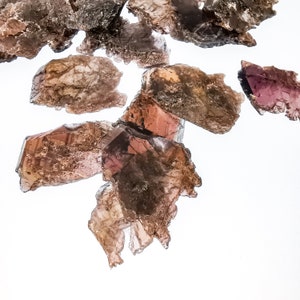 Puede incluir: Una colección de cristales de mica marrones y morados con una apariencia estratificada y translúcida. Los cristales están dispuestos en un grupo sobre un fondo blanco.