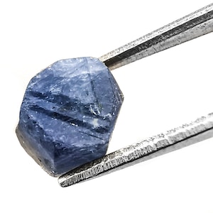 Puede incluir: Primer plano de una gema de zafiro azul sostenida por unas pinzas. El zafiro tiene una forma facetada, geométrica y un color azul intenso.