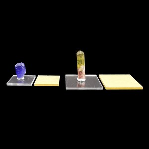 Puede incluir: Tres soportes de exhibición de acrílico transparente con tres cristales de diferentes colores. Los cristales son un azul, un rosa y verde, y un amarillo y verde. Los soportes están sobre un fondo negro.