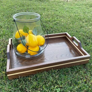 May include: A wooden serving tray with a dark brown finish and a light wood accent, holding a clear glass jar filled with yellow lemons and green leaves. The jar has a black metal handle. The tray is on green grass.