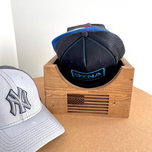 May include: A wooden hat holder with an engraved American flag design, holding several baseball caps. One cap is gray with a white logo, and another is black with a blue and teal design. The holder is made of light brown wood.