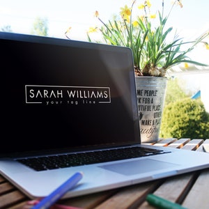 May include: A silver laptop computer with a black screen displaying a logo that reads "SARAH WILLIAMS your tag line". The laptop is sitting on a wooden table with a potted plant and a pencil in the foreground.