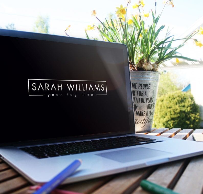 May include: A silver laptop computer with a black screen displaying a logo that reads "SARAH WILLIAMS your tag line". The laptop is sitting on a wooden table with a potted plant and a pencil in the foreground.