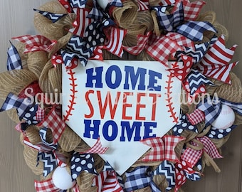 May include: A baseball-themed wreath with a burlap base and red, white, and blue ribbon. The wreath features a white home plate sign with the text "Home Sweet Home" in blue and red lettering. The wreath is decorated with baseballs, polka dot and chevron patterned ribbons, and gingham check.