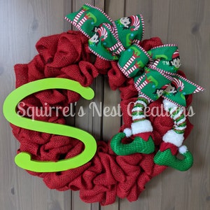 May include: A red burlap Christmas wreath with a green and white striped bow featuring elf illustrations. A large green letter "S" is attached to the wreath. Two green elf legs with white boots are hanging from the bow.