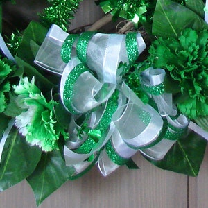 May include: A green and white wreath with a large green and white bow. The wreath is decorated with green carnations and green and white tinsel.