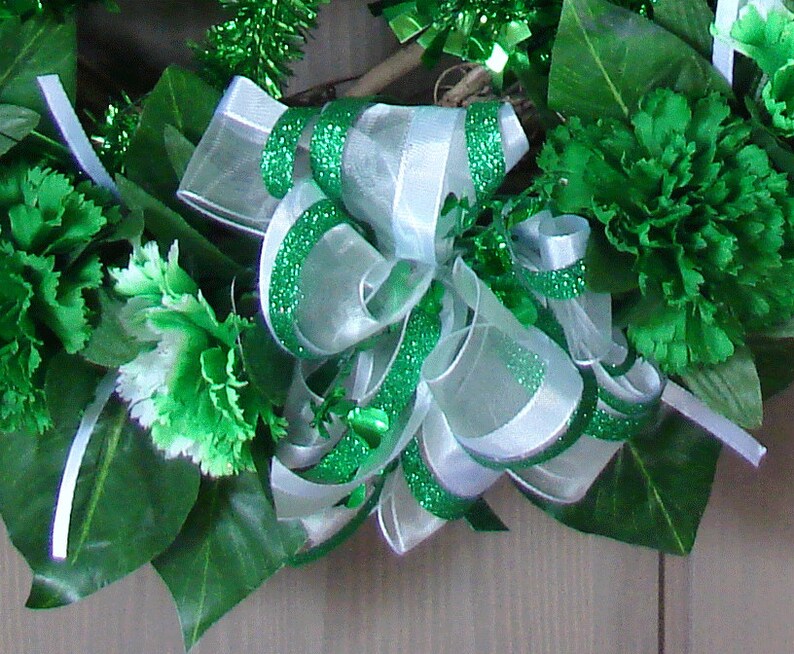 May include: A green and white wreath with a large green and white bow. The wreath is decorated with green carnations and green and white tinsel.