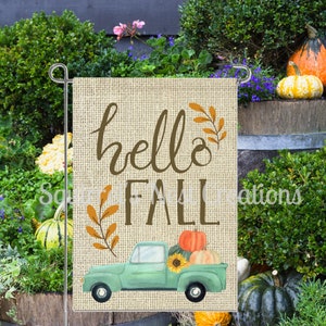 May include: A burlap garden flag with the text "hello fall" in brown script. A green pickup truck with a sunflower and pumpkins in the bed is driving on a dirt road.