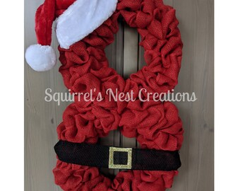 May include: Red burlap wreath shaped like Santa Claus with a white faux fur hat and a black belt with a gold buckle. The wreath says "Squirrel's Nest Creations".