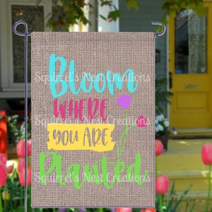 May include: A burlap garden flag with the text "Bloom Where You Are Planted" in colorful letters. The flag has a floral design with pink, purple, and yellow flowers.