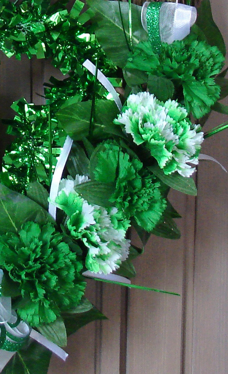 May include: A green and white floral wreath with green leaves and white ribbon. The wreath is decorated with green and white carnations.