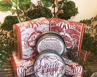 May include: Two round metal tins with red and white labels that say "Patchouli" and "Patchouli Conditioner". The tins are sitting on top of two brown bars of soap with red and white labels that say "Patchouli Shampoo".