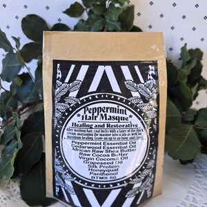 Op de afbeelding: Een bruine papieren zak met een zwart-wit etiket waarop "Peppermint Hair Masque" staat en de ingredi&euml;nten worden vermeld: pepermuntolie, cederhouten olie, Afrikaanse ruwe sheaboter, ruwe cacaoboter, virgin kokosolie, druivenpitolie, zijdeprote&iuml;ne, honingwas, panthenol, BTMS 50.