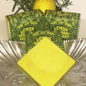 May include: A yellow bar of soap with the text "Lemon Rosemary Soap" on the wrapper. The soap is sitting on a silver tray with a lemon and rosemary sprig in the background.