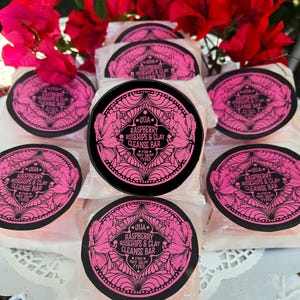May include: Several round, wrapped bars of soap with black and pink labels. The labels read "Raspberry Rosehips & Clay Cleanse Bar" and "For Dry Skin Types." Red flowers are in the background.