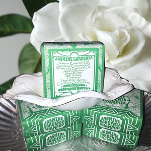 Puede incluir: Cuatro barras de champ&uacute; verde y blanco etiquetadas como "Champ&uacute; de Jazm&iacute;n y Gardenia". Las barras est&aacute;n apiladas una encima de la otra en un cuenco plateado. El cuenco est&aacute; rodeado por una flor blanca.