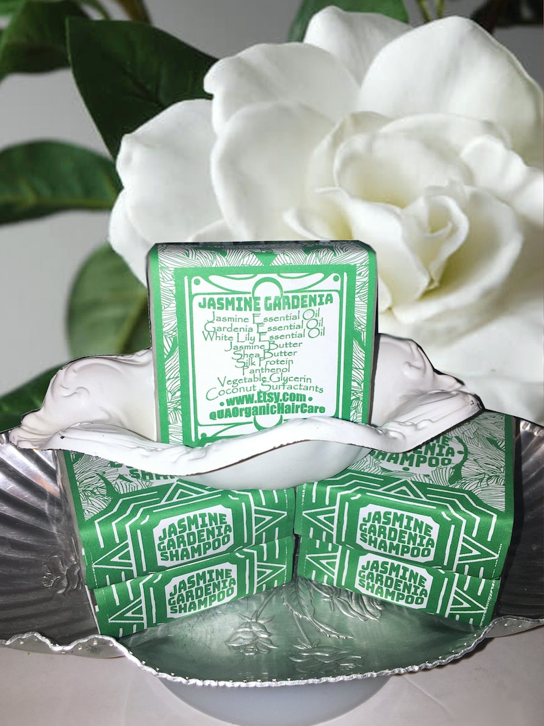 Puede incluir: Cuatro barras de champ&uacute; verde y blanco etiquetadas como "Champ&uacute; de Jazm&iacute;n y Gardenia". Las barras est&aacute;n apiladas una encima de la otra en un cuenco plateado. El cuenco est&aacute; rodeado por una flor blanca.