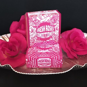 May include: Two pink and white bars of soap with the text "Fresh Roses Conditioner" on them. The soap bars are on a silver tray with pink rose petals.
